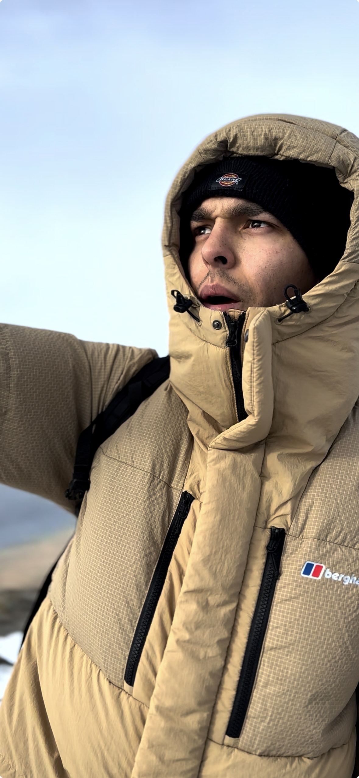 A man in a tan hooded puffer jacket and black beanie looks off to the side.