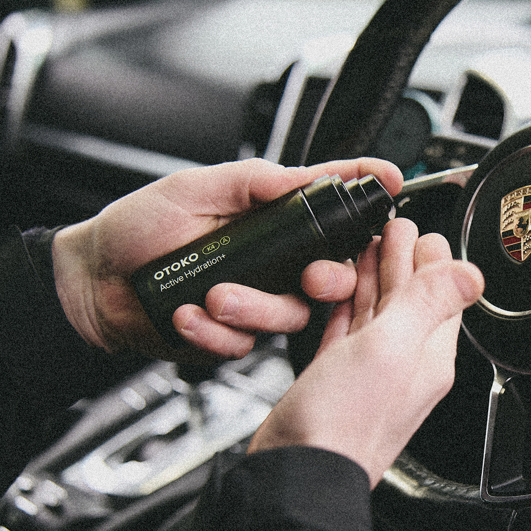 Hands holding a bottle of 'OTOKO Active Hydration+' near a car steering wheel.