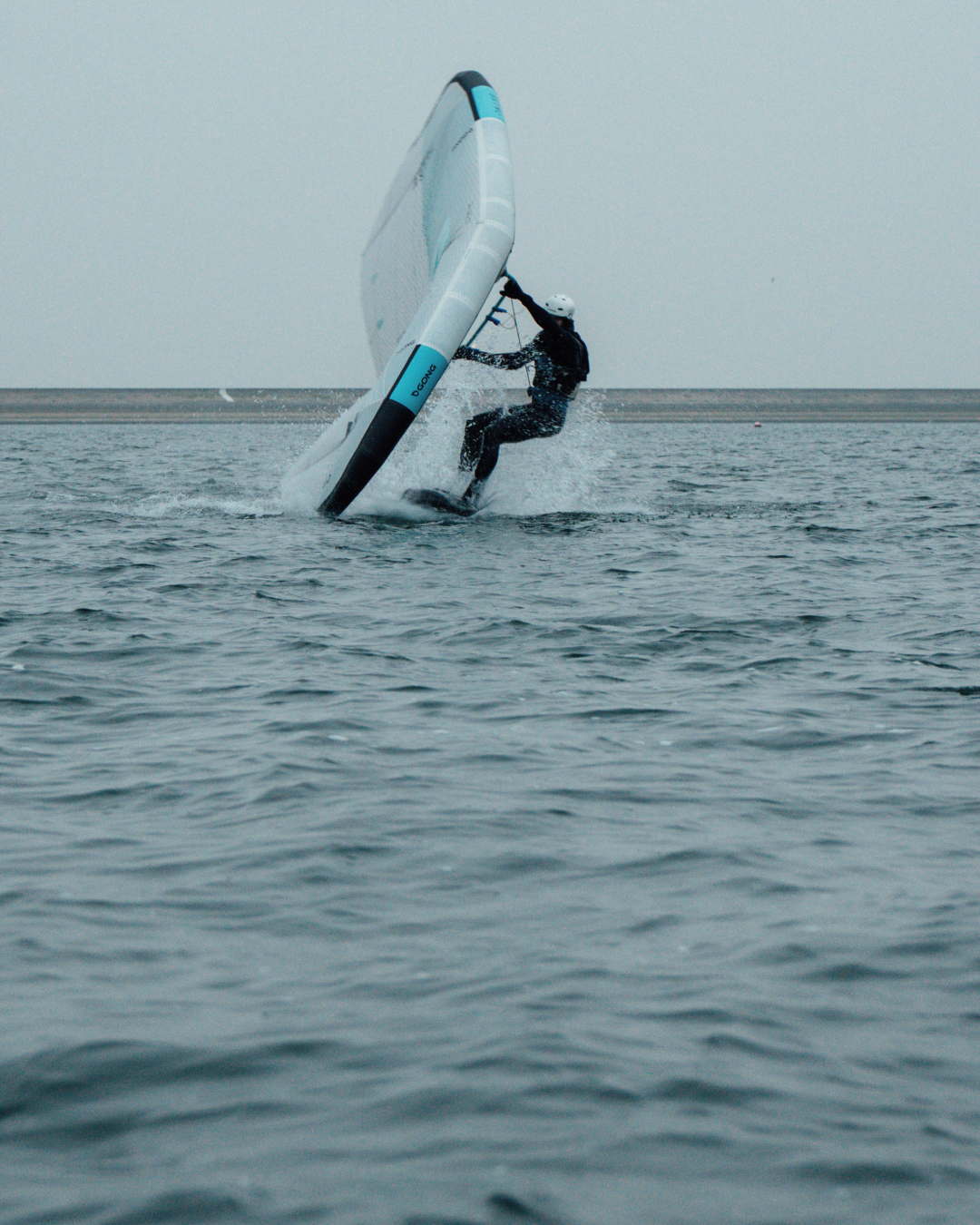 A person in a wetsuit wing foils across the water, making a sharp turn and creating a splash.