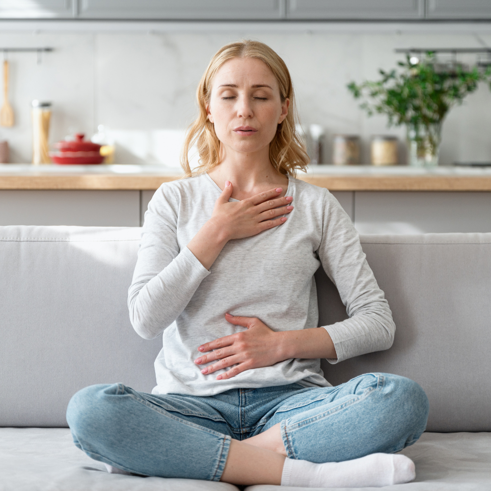 A woman sits cross-legged on a couch with her eyes closed, one hand on her chest and the other on her stomach.