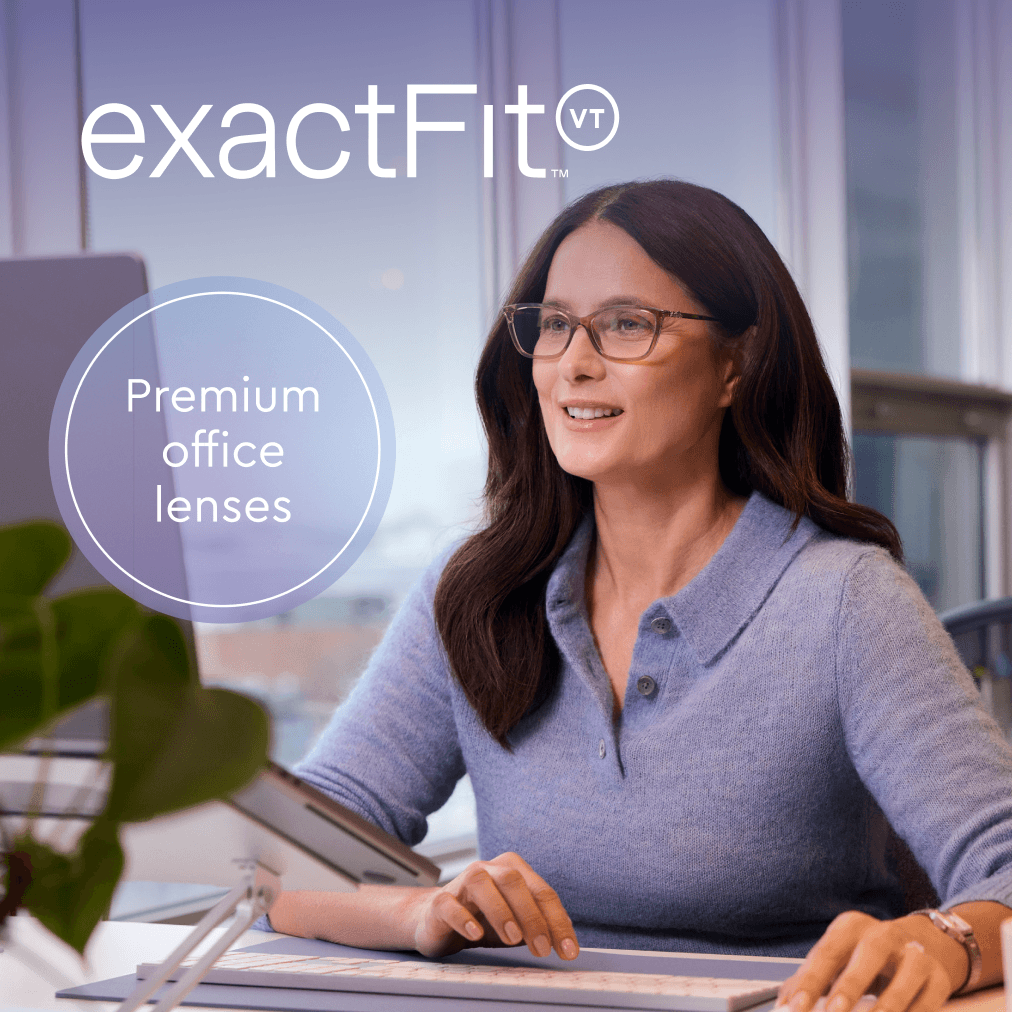 A smiling woman with glasses sits at her desk in an office, using a computer.