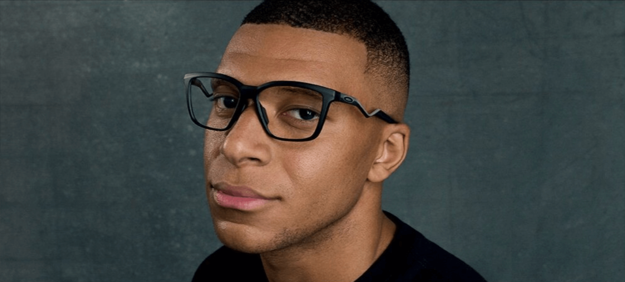 Person wearing black glasses, looking at the camera against a dark background.