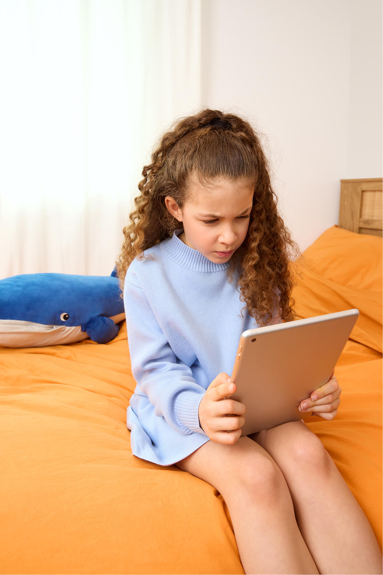 Child sitting on bed with tablet, blue plush toy in background.