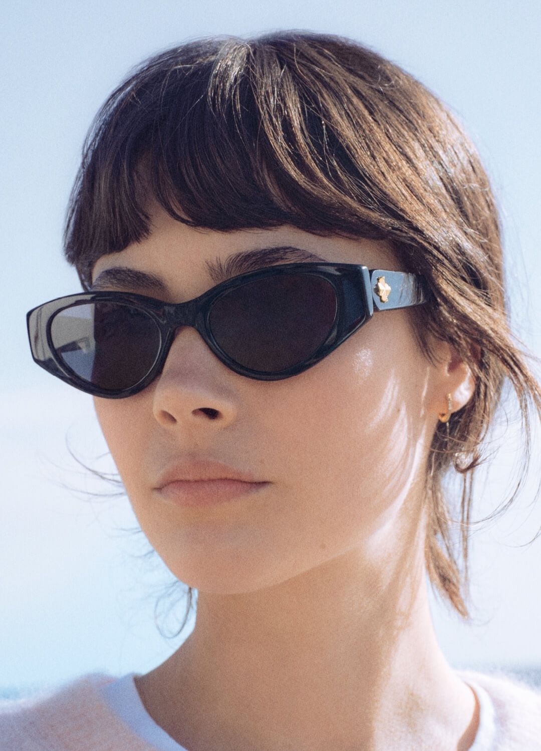 A close-up of a woman with brown hair and bangs wearing black cat-eye sunglasses with a gold emblem.