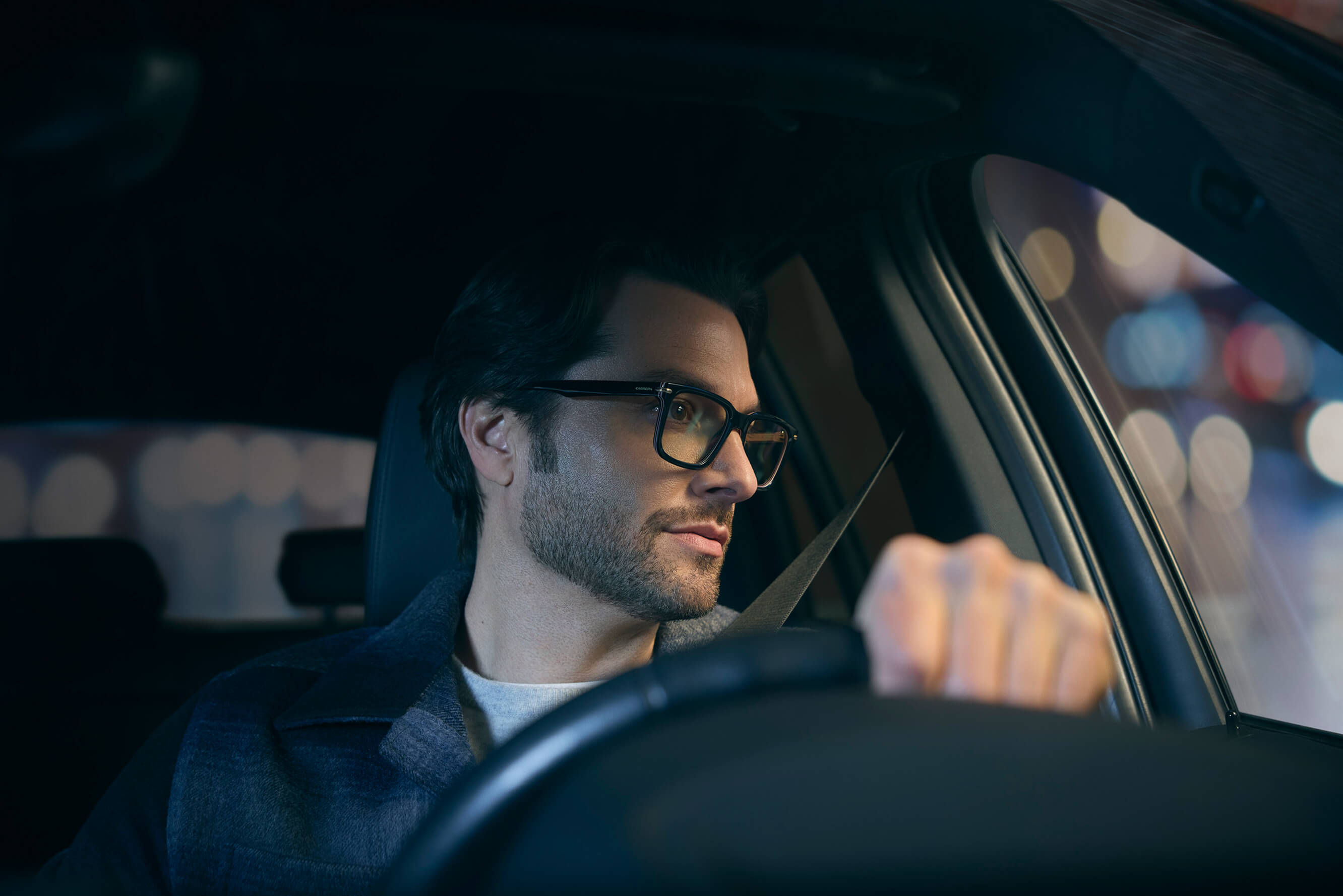 A man with glasses and a beard drives a car at night with city lights in the background.
