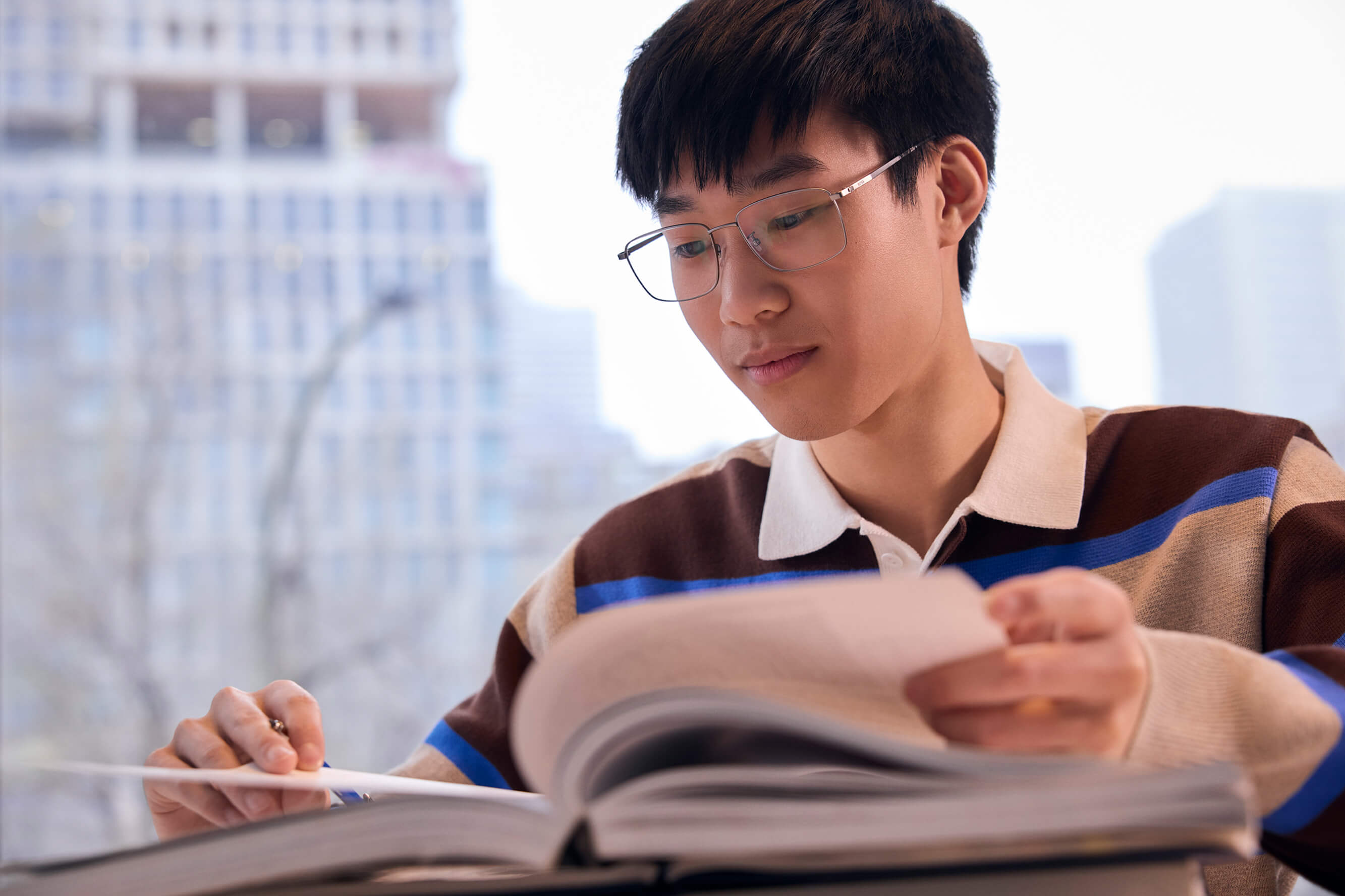 A young man with glasses and a striped shirt sits indoors reading a book by a window.