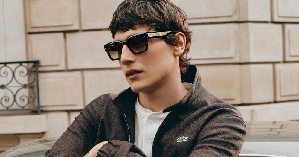 A man with shaggy brown hair wears tortoiseshell sunglasses and a brown Lacoste jacket outdoors.