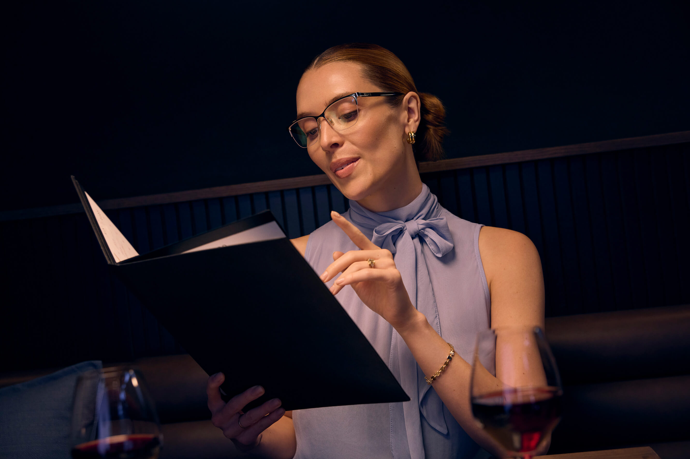 A woman wearing glasses sits at a restaurant table, looking at a menu and pointing to an item.