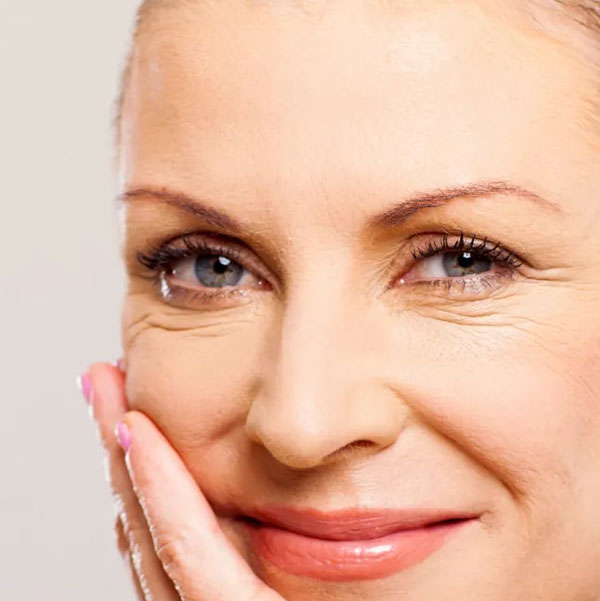 A close-up of a smiling woman with wrinkles around her eyes, touching her cheek with her hand.