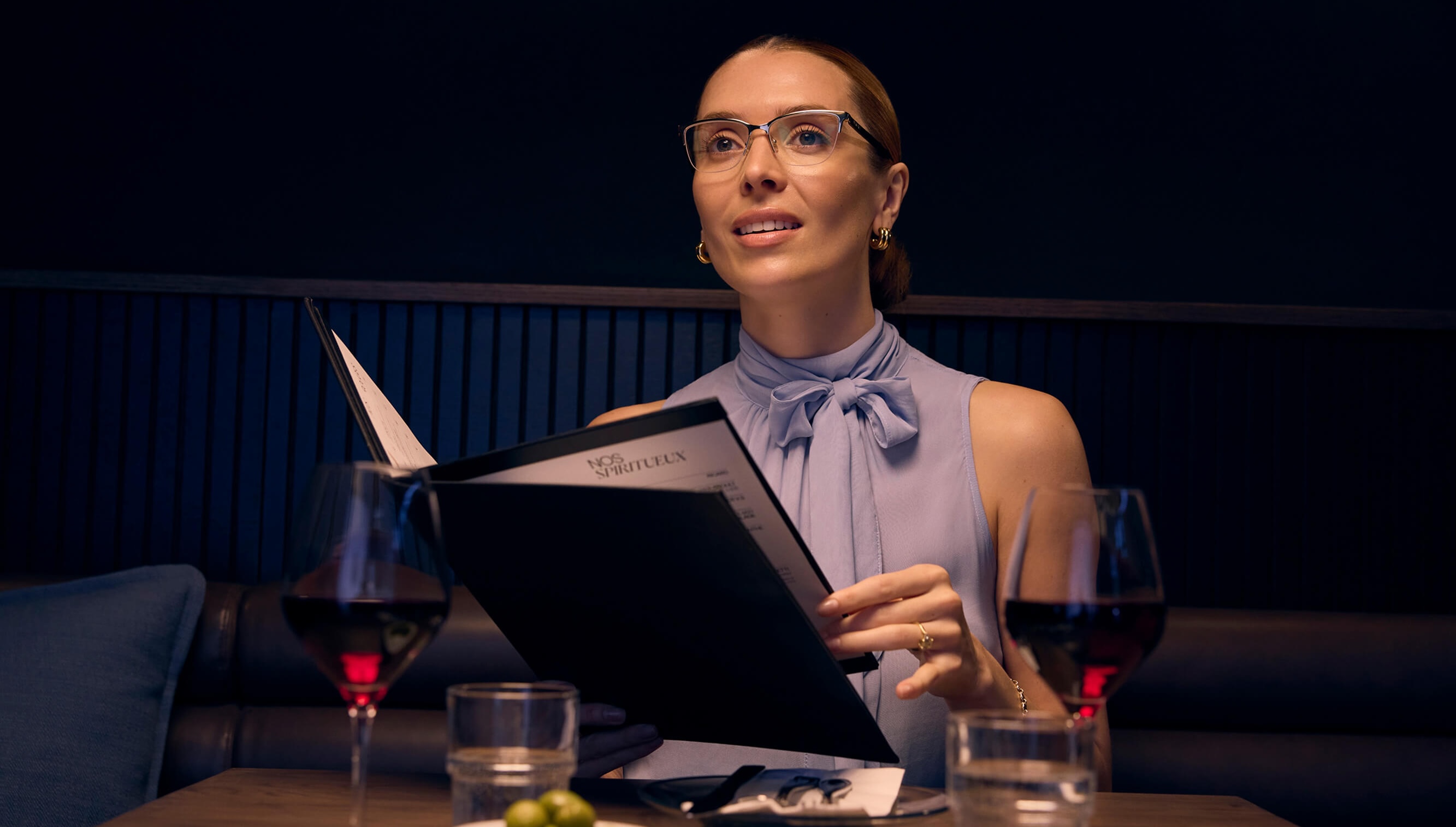A woman with progressive lenses holds a menu while sitting at a restaurant table with glasses of wine.