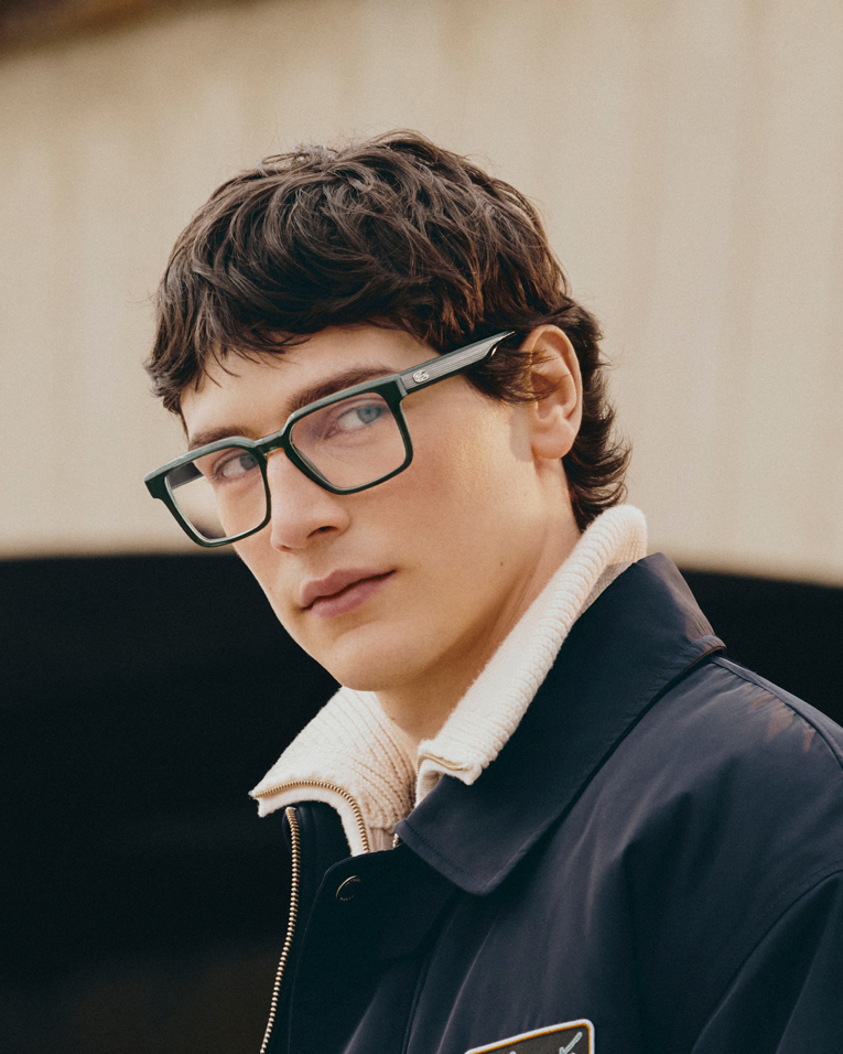 A young man with brown hair and green rectangular glasses looks over his shoulder.