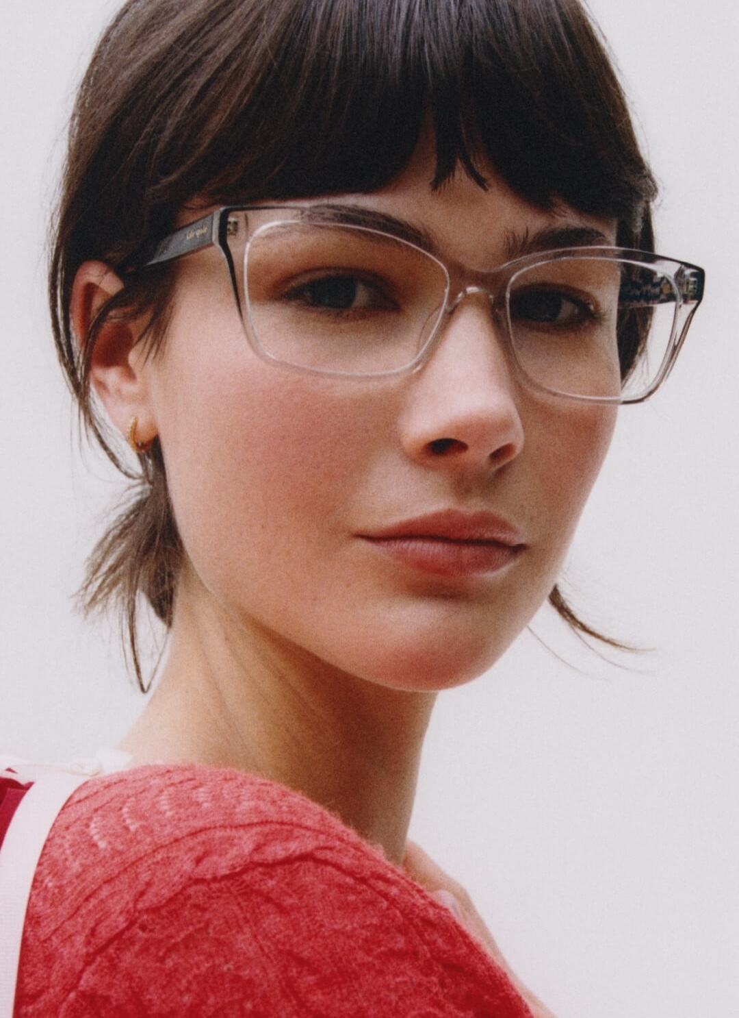 A close-up of a woman with short dark hair and clear-framed eyeglasses, wearing a red knit sweater.