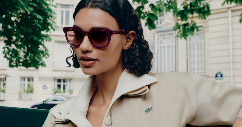 A woman with dark curly hair wearing Lacoste burgundy sunglasses and a beige Lacoste jacket outdoors.