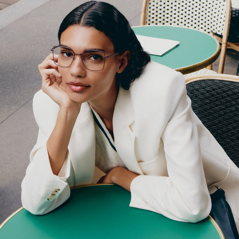 A woman wearing glasses and a white blazer rests her chin on her hand at a green cafe table.