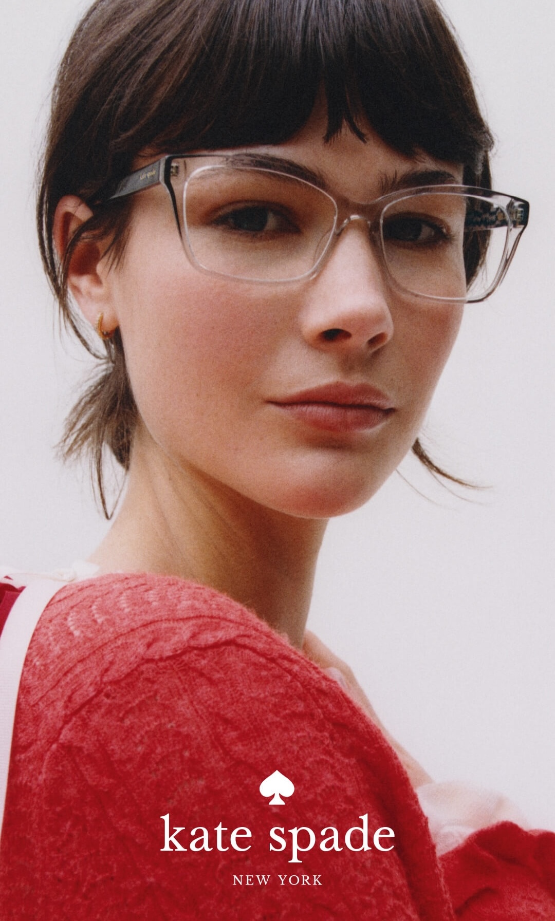 A woman with short dark hair and clear-framed eyeglasses wears a red sweater, looking toward the camera.