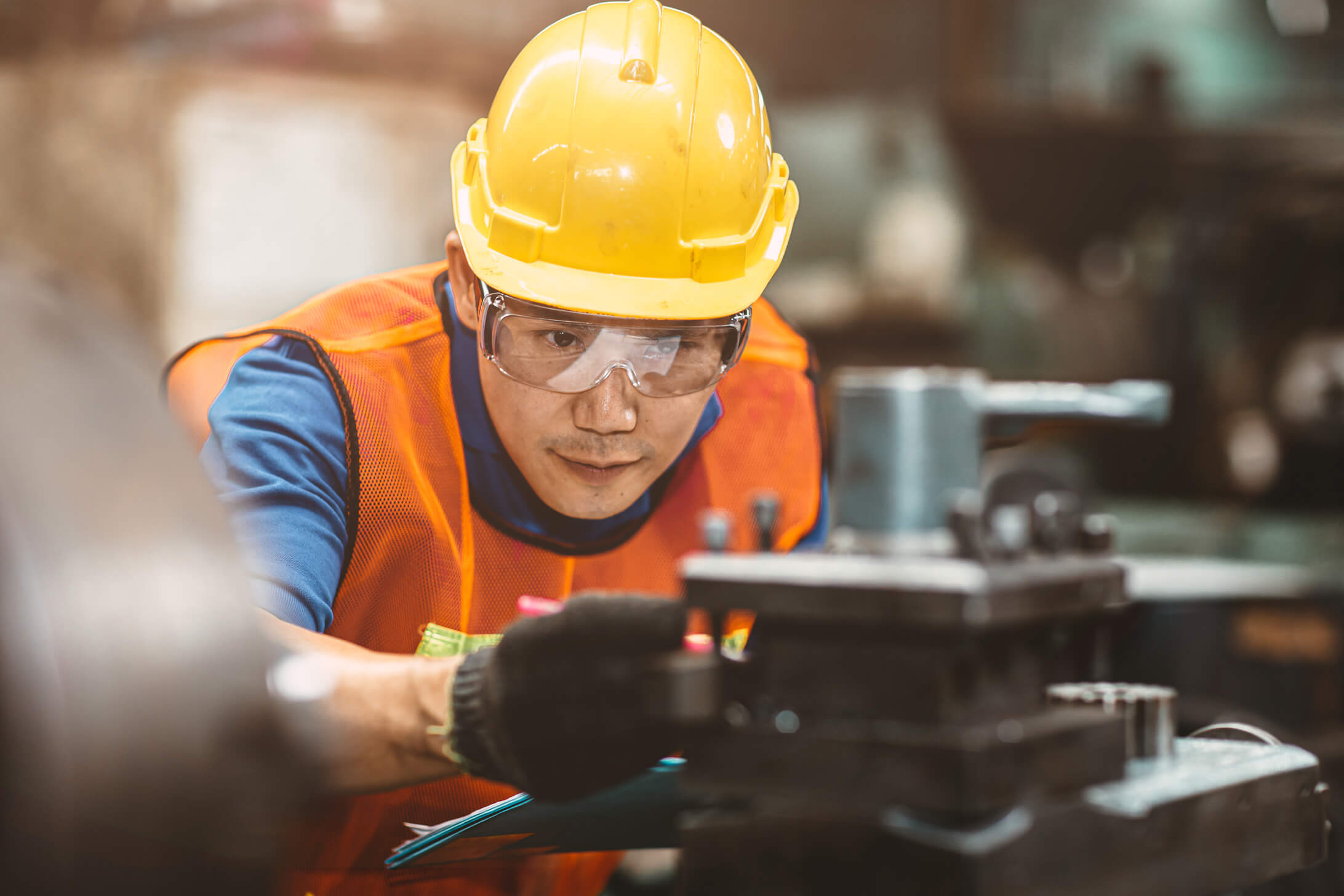 Un ouvrier d’usine portant un casque de sécurité jaune, un gilet de sécurité et des lunettes de protection examine attentivement une machine.