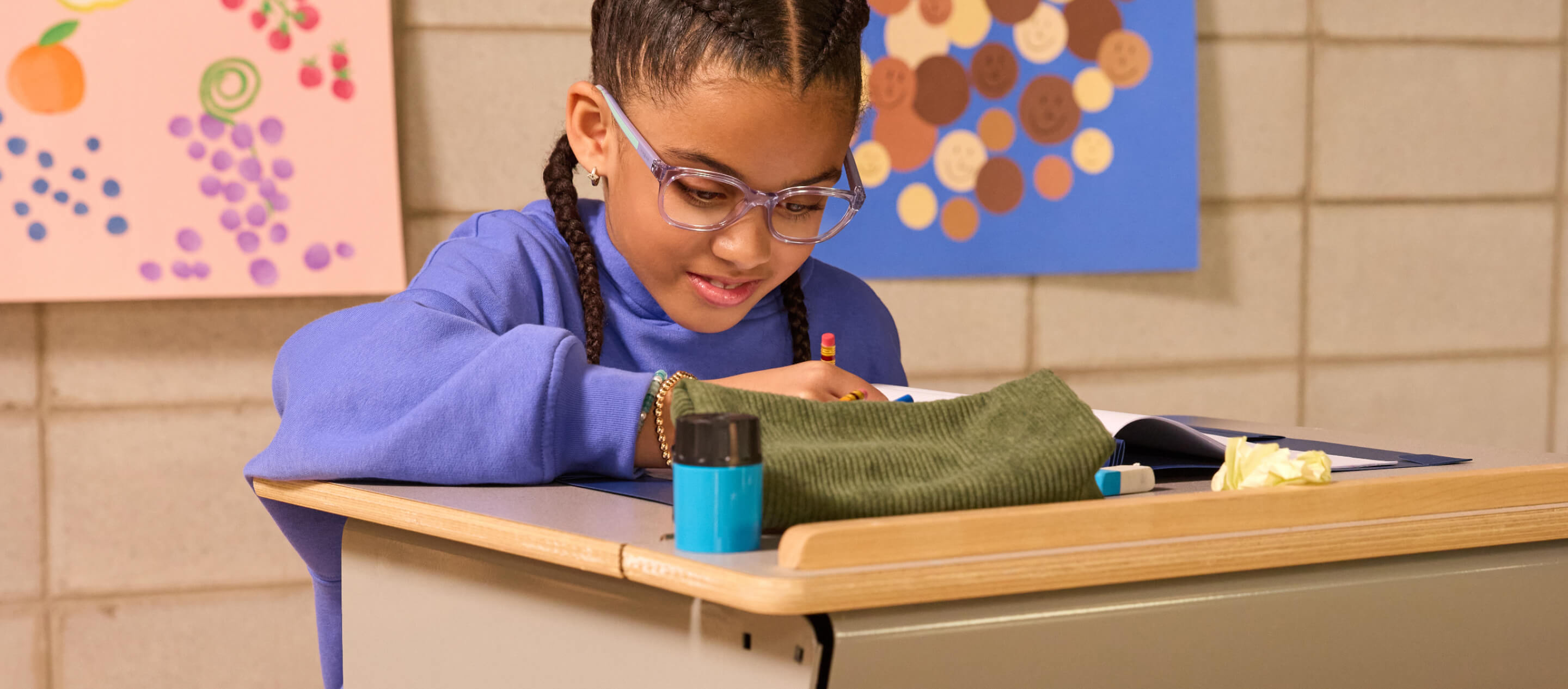 A young girl with glasses and braided hair sits at a school desk, writing in a notebook.