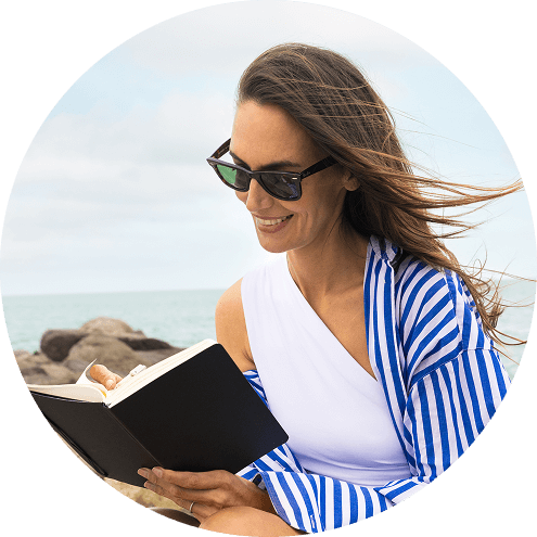Une femme souriante portant des lunettes de soleil lit un livre au bord de la mer, les cheveux au vent.