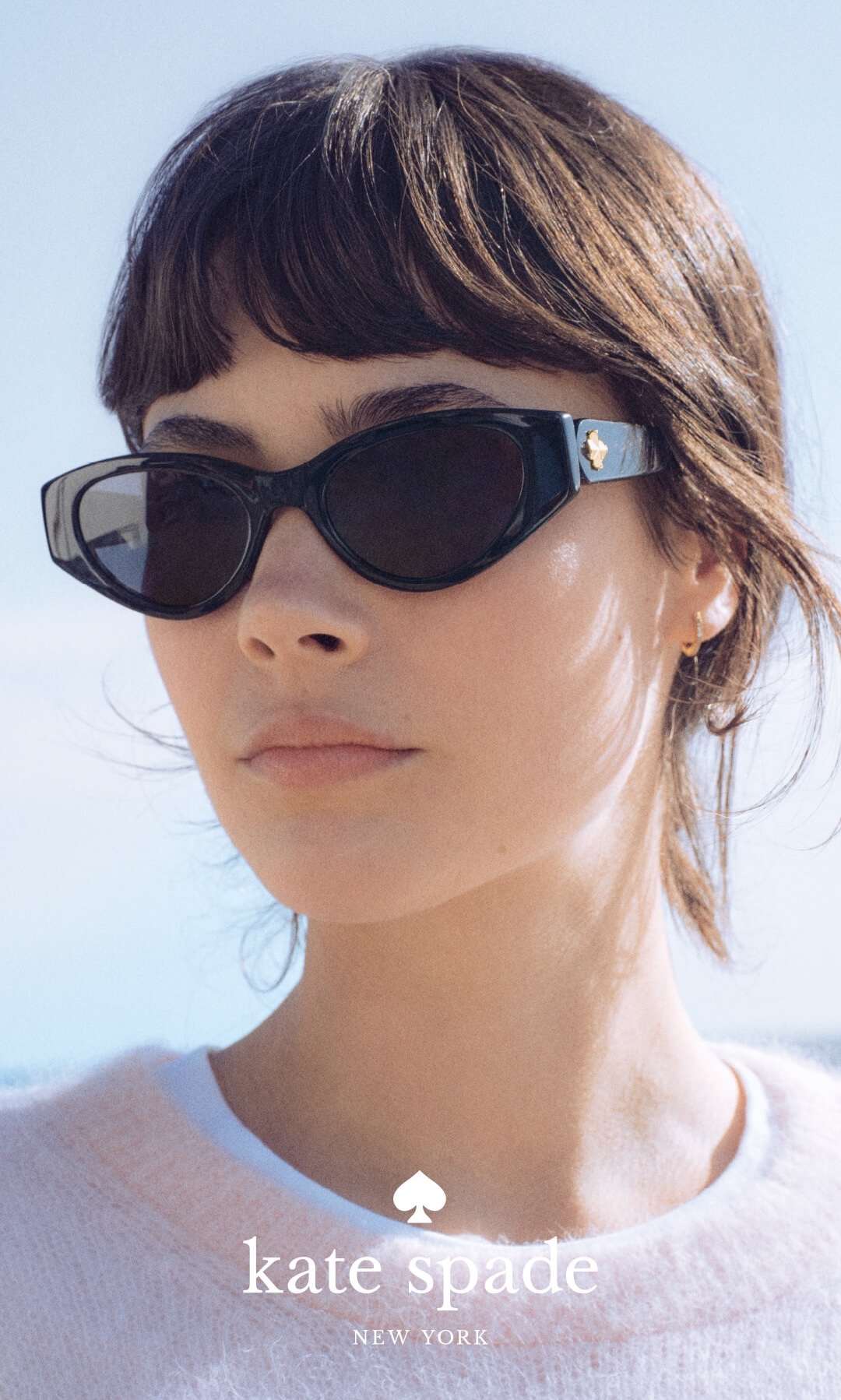 A close-up of a woman with brown hair and bangs wearing black sunglasses with a gold spade detail.