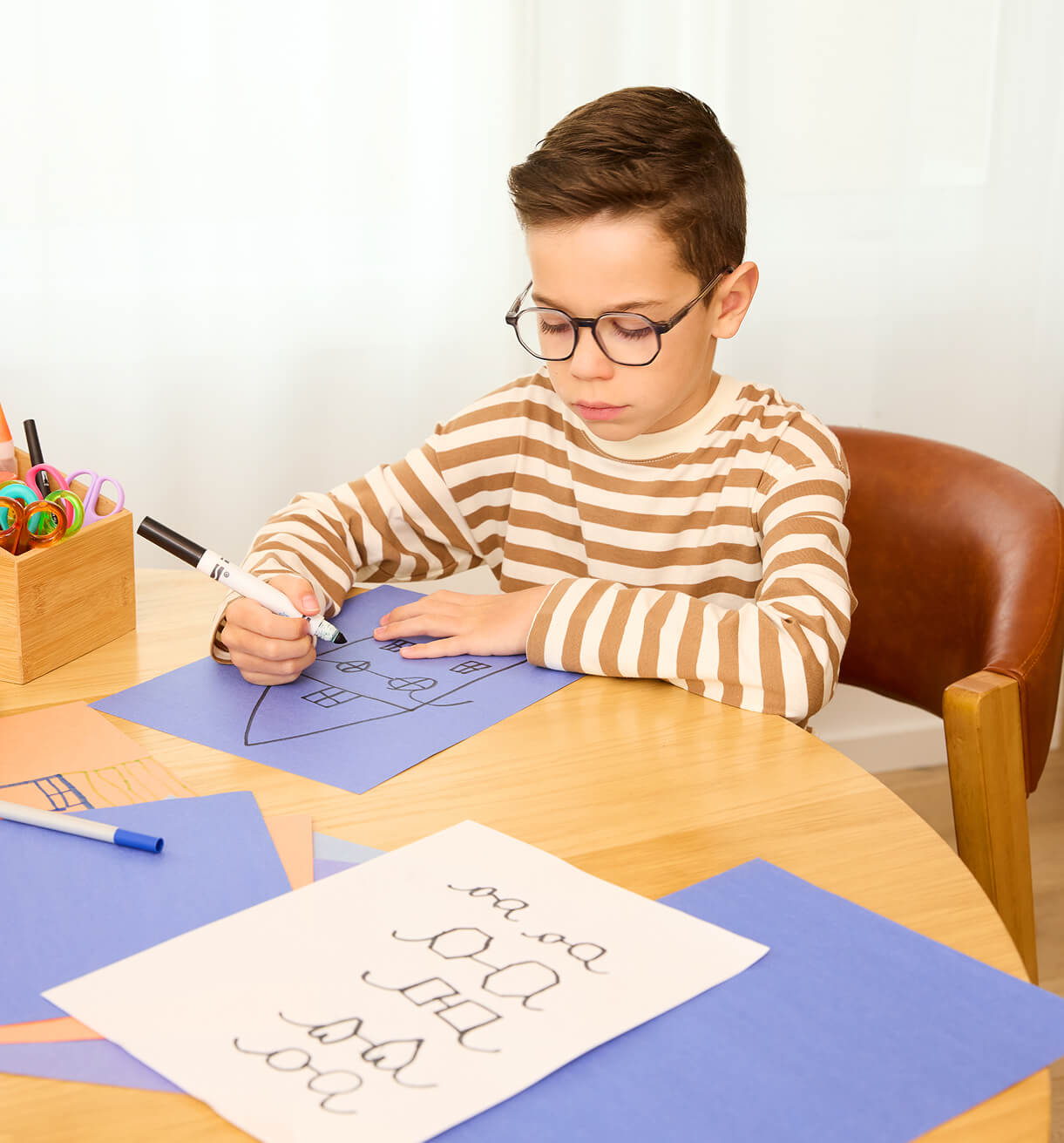Child wearing glasses drawing on blue paper at a table with art supplies.