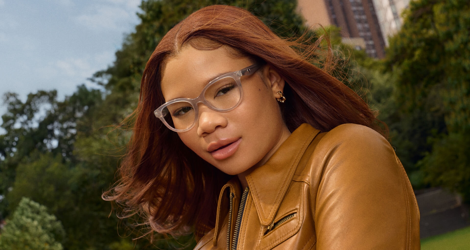 Storm Reid with reddish-brown hair and Coach, clear-framed glasses wearing a tan leather jacket looks at the camera.