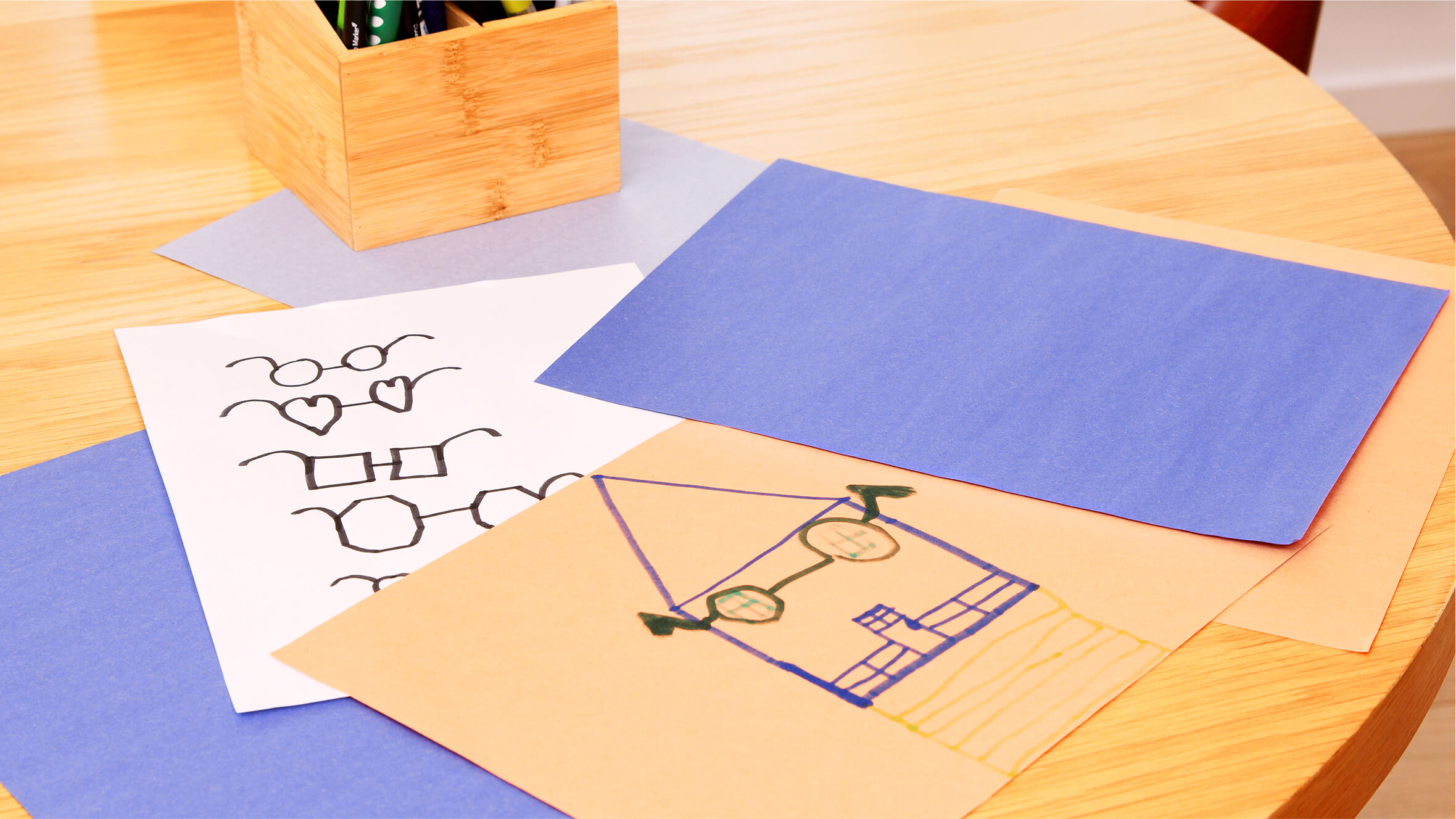 Drawings of glasses and a house on colored paper on a wooden table.