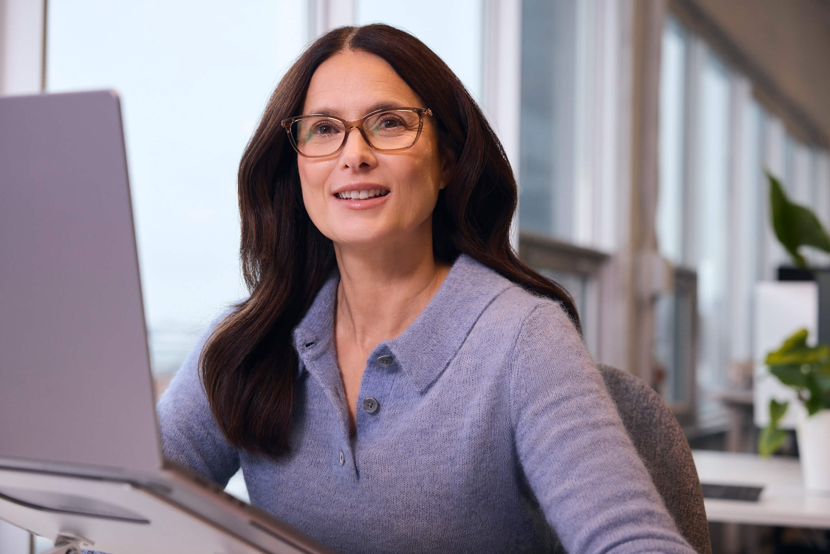A woman with glasses and dark hair smiles while sitting in front of a laptop in an office.