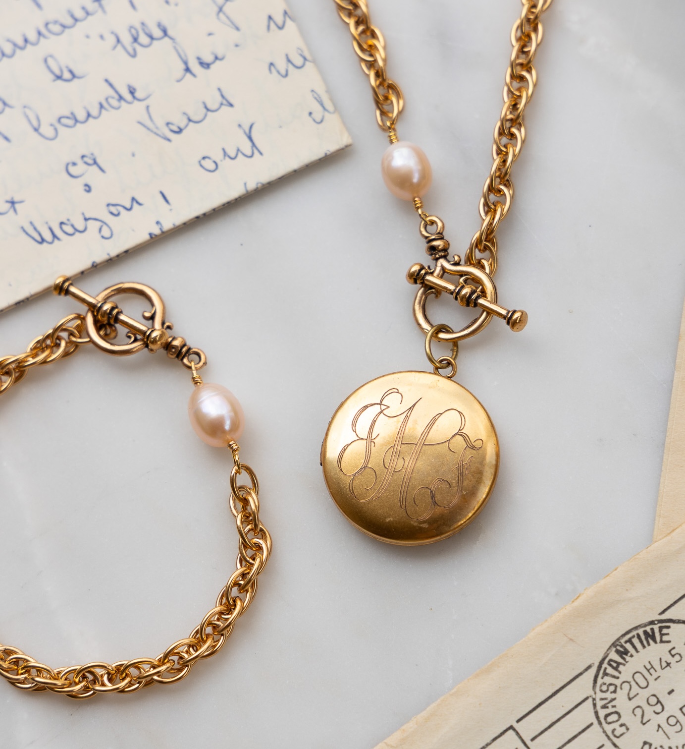 A gold engraved locket necklace and matching bracelet with pearls, lying on a marble surface with letters.