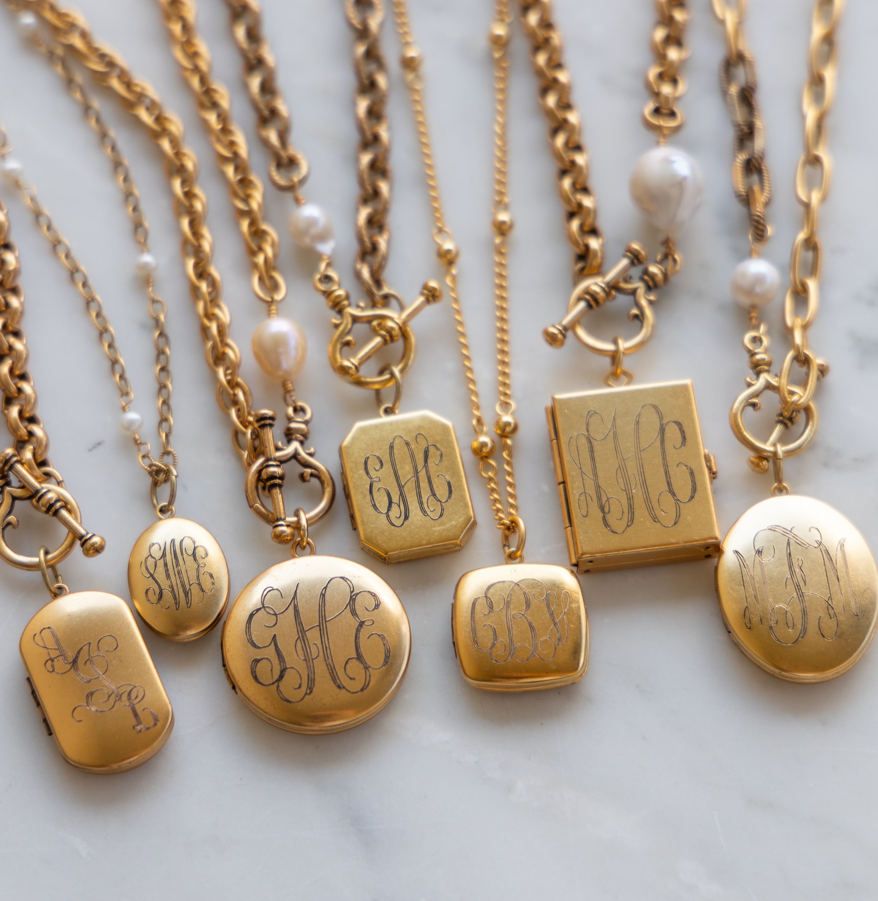 A collection of vintage gold locket necklaces with monograms on a marble surface.
