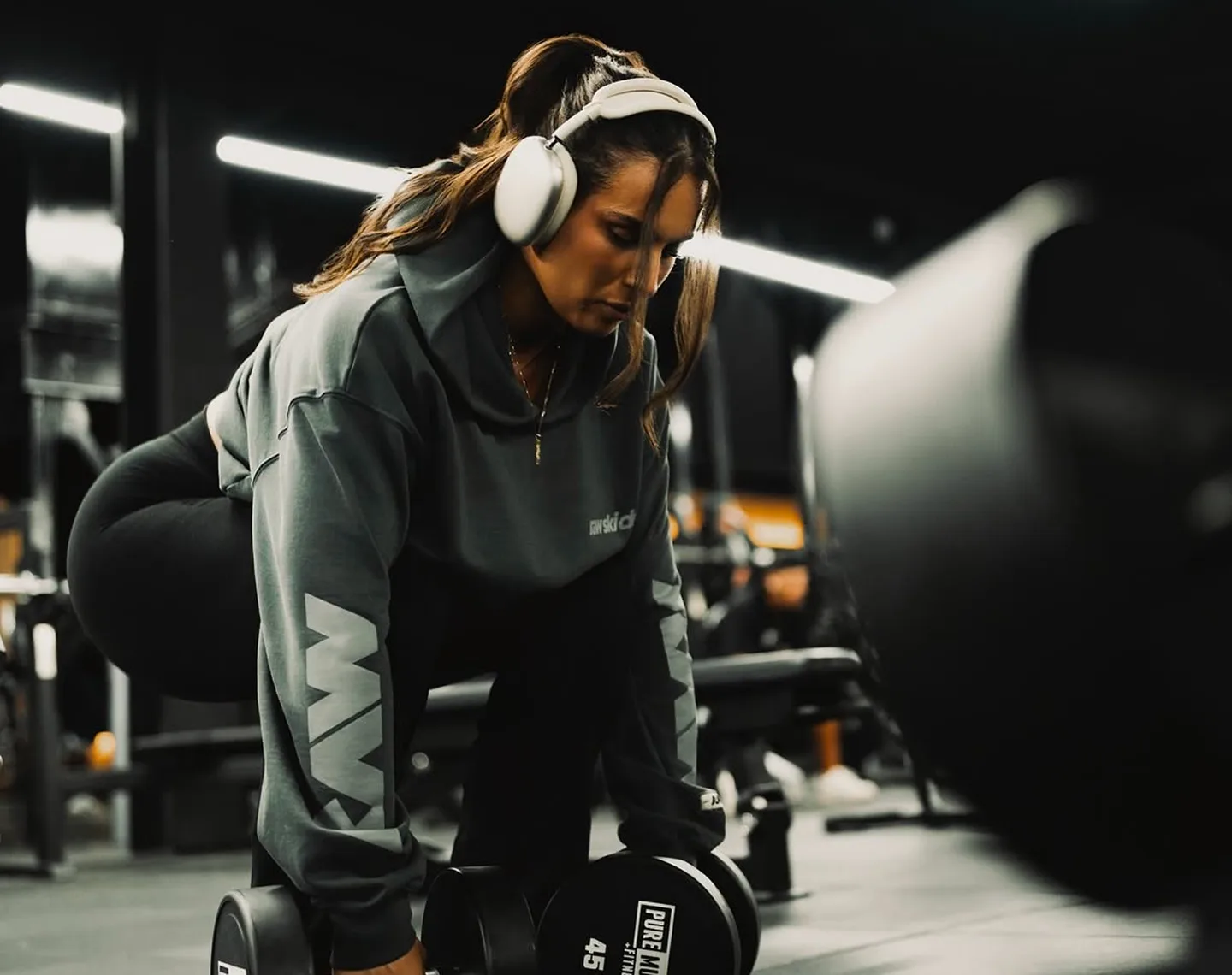 A woman wearing headphones and a grey hoodie bends down to pick up a dumbbell in a gym.