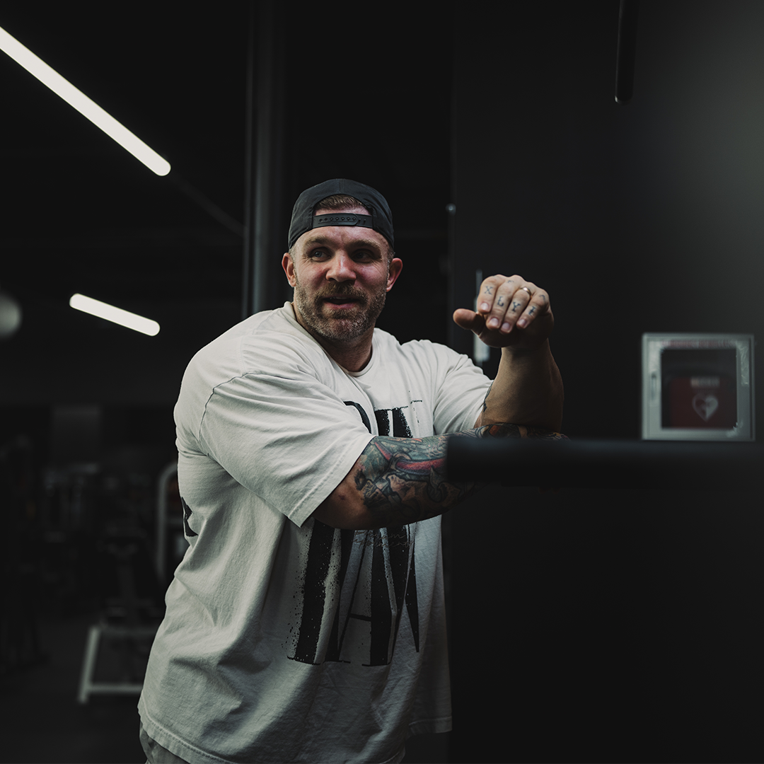 Man in a gym leaning on equipment, wearing a cap and white shirt.