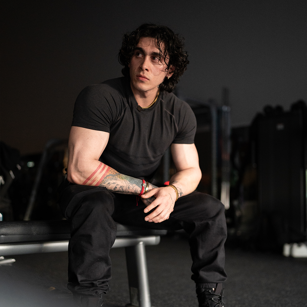 Man with tattoos sitting on a bench in a gym, looking to the side.