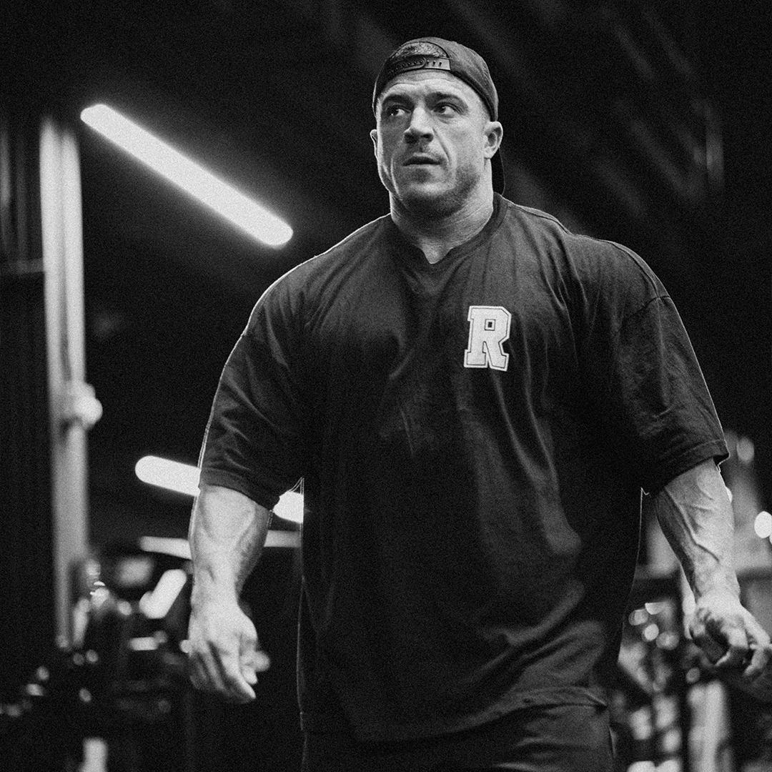 Black and white photo of a muscular man in a gym, wearing a cap and a shirt with 'R'.