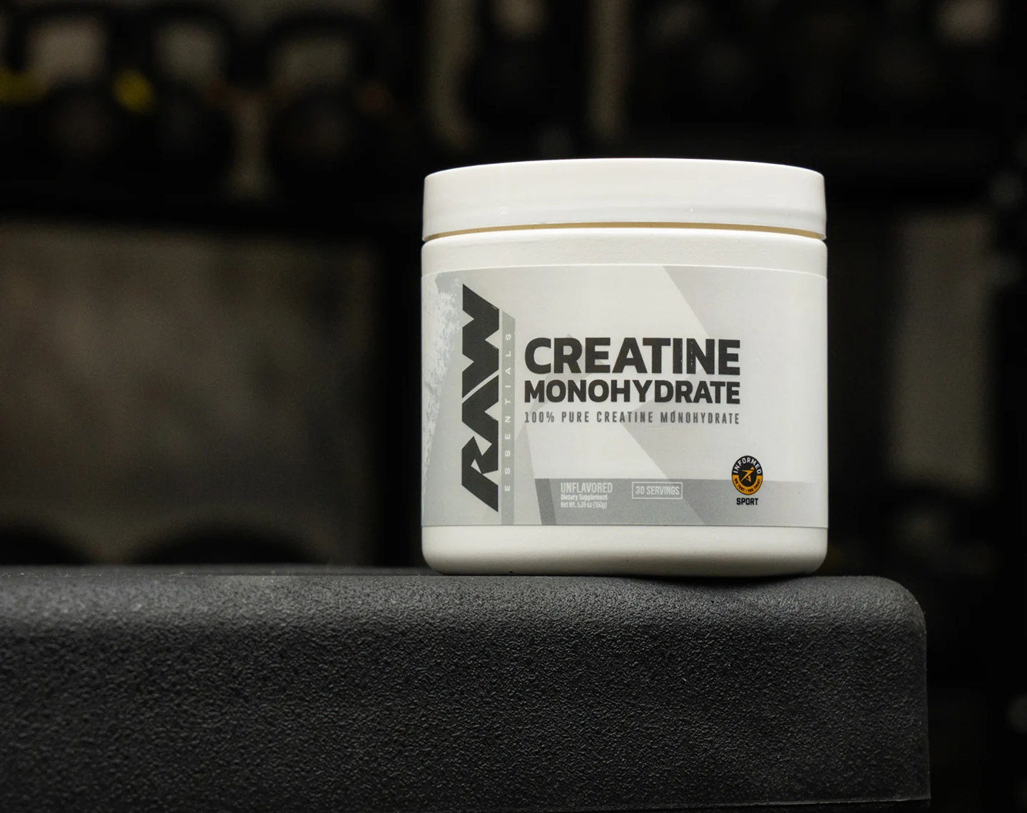A white tub of Raw Essentials Creatine Monohydrate supplement resting on a black surface in a gym.