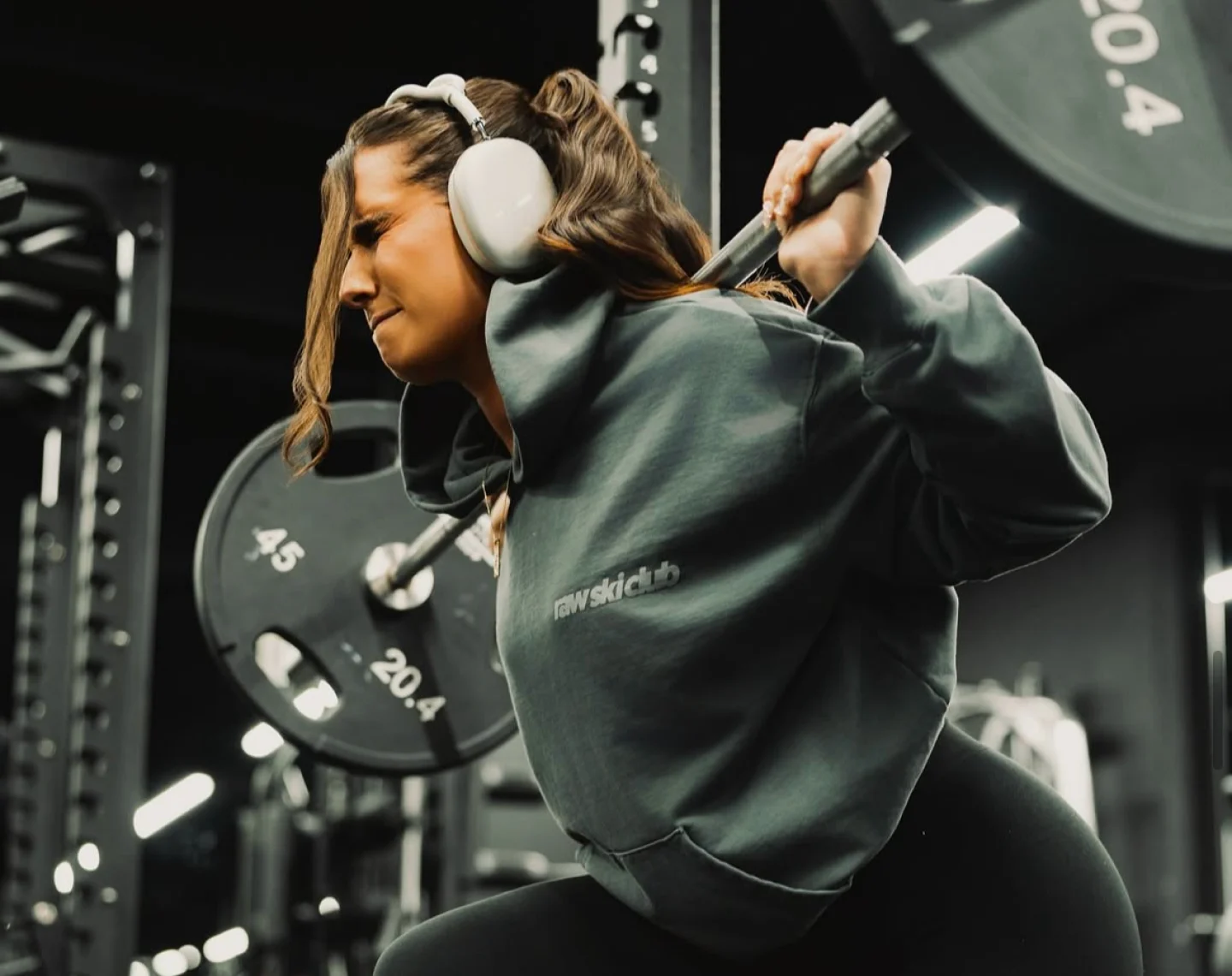 A woman in a grey hoodie and headphones performs a barbell squat in a gym.