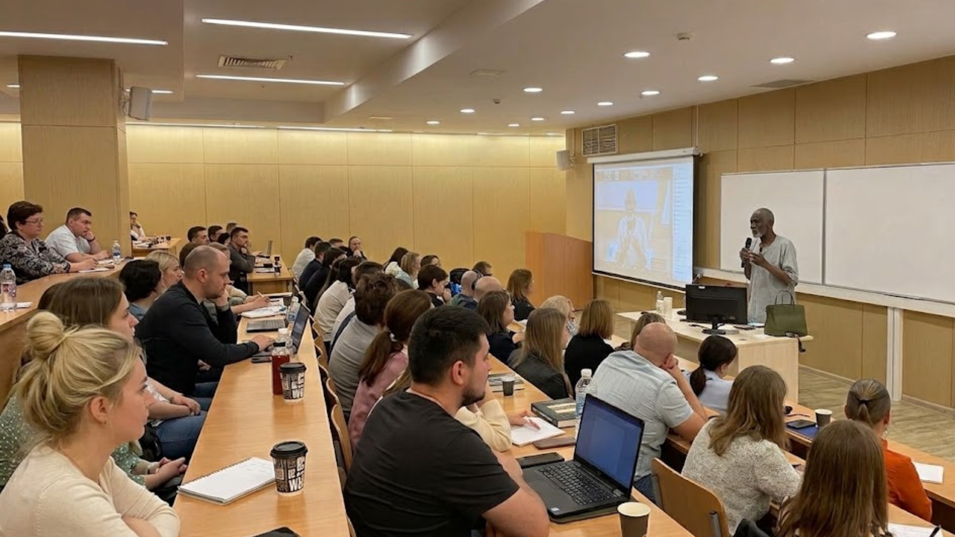 A man lectures to an audience seated in a tiered classroom or lecture hall.