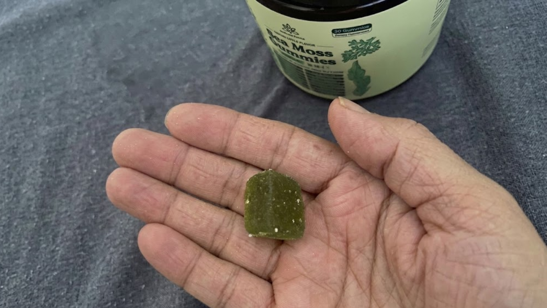 A close-up of a hand holding a single green sea moss gummy with the bottle in the background.