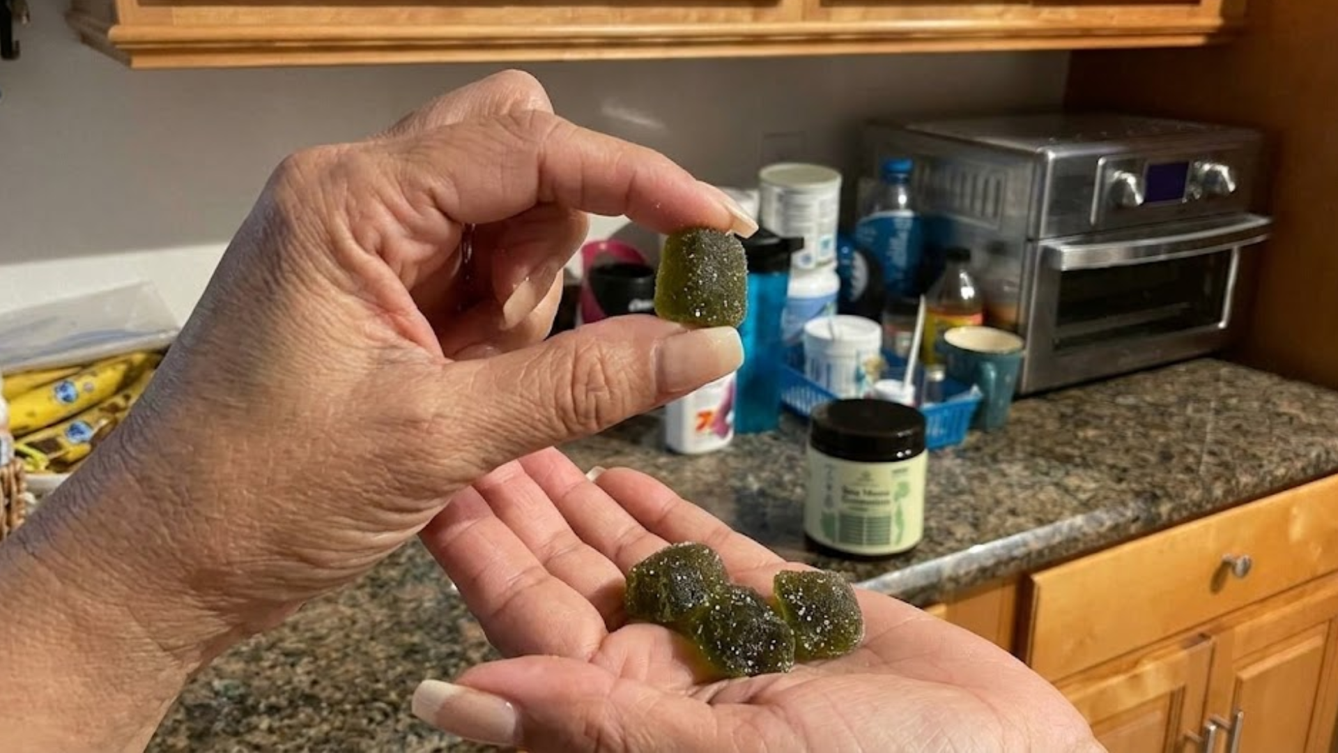A close-up of a person's hands holding several dark green, sugar-coated gummies in a kitchen.