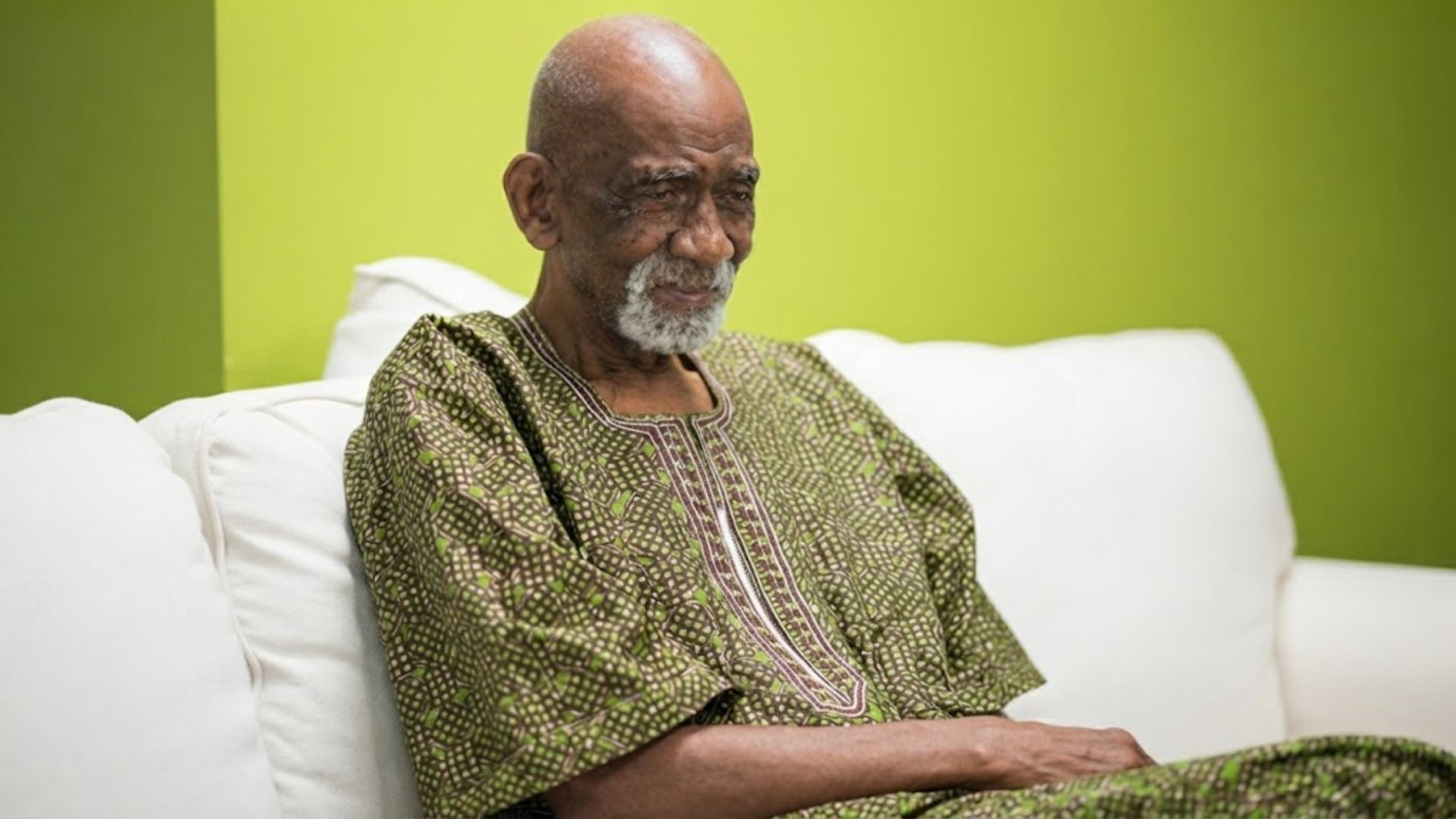 An elderly Black man with a white goatee, wearing a patterned green tunic, sits on a white couch.