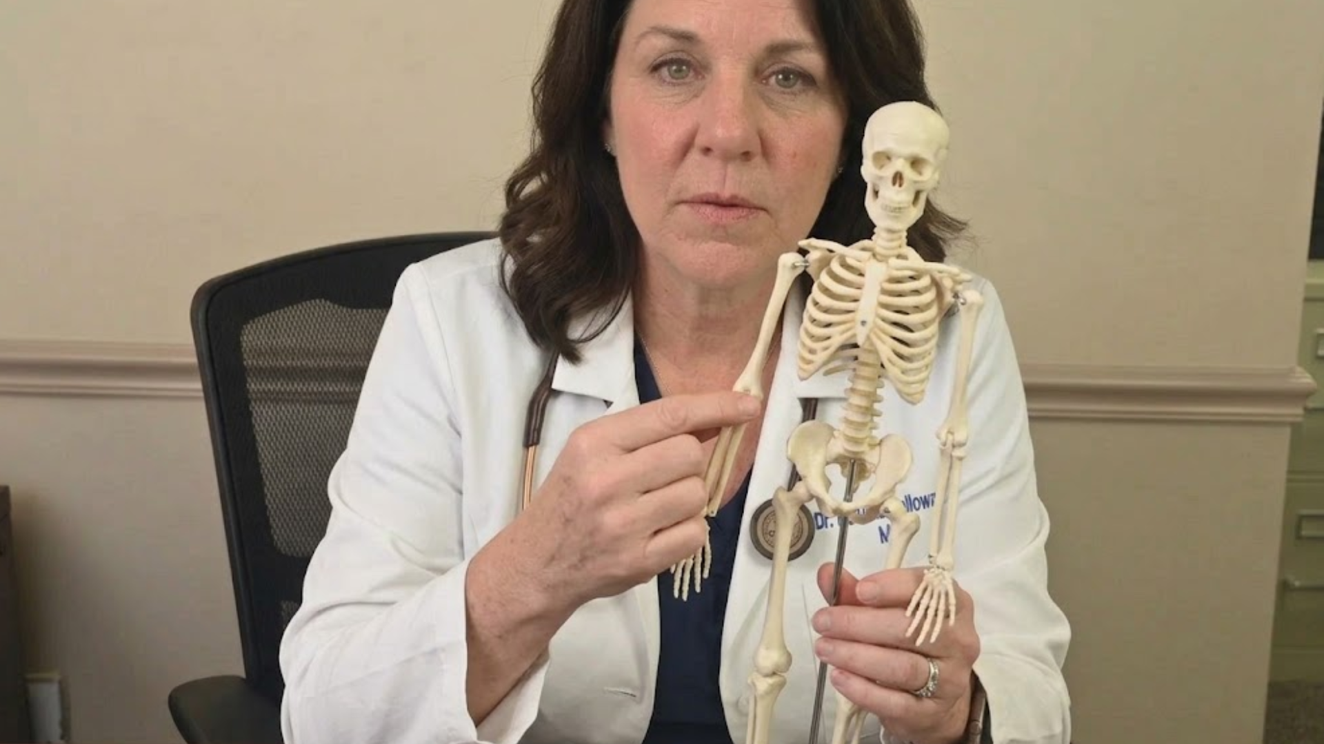 A doctor in a white coat points to the arm of a small anatomical skeleton model.