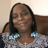 A woman with braided hair adorned with blue and white beads smiles with her eyes closed.