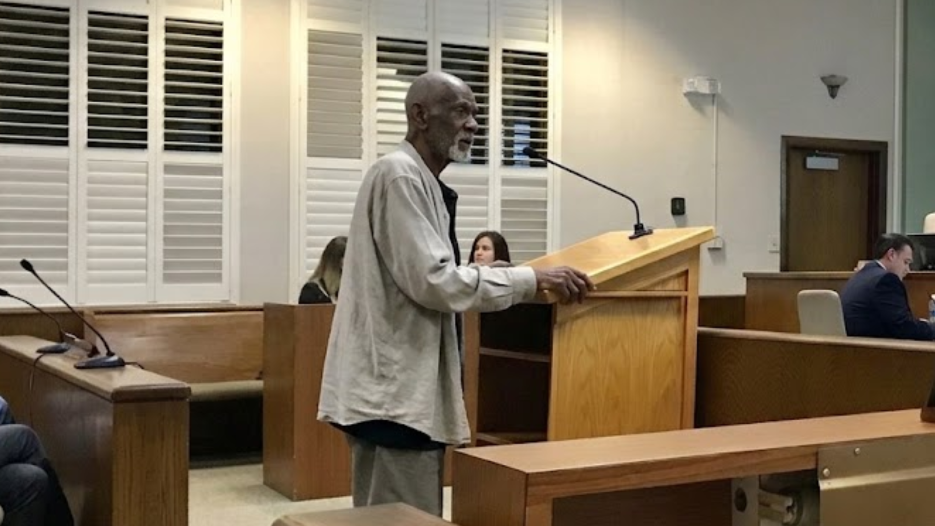 An elderly Black man stands at a wooden lectern in what appears to be a courtroom or hearing room.