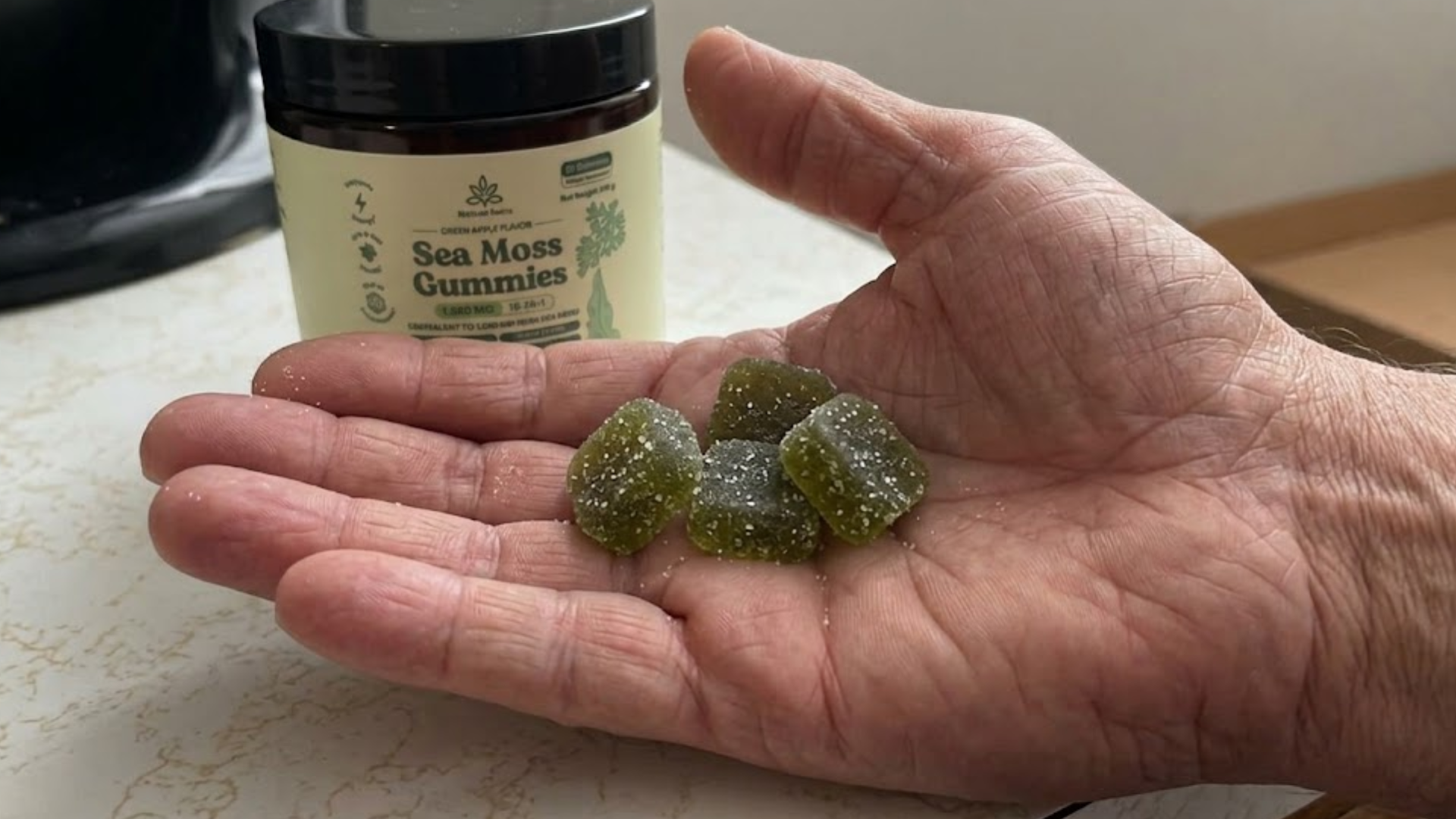 A hand holding several green, sugar-coated sea moss gummies with the product jar in the background.