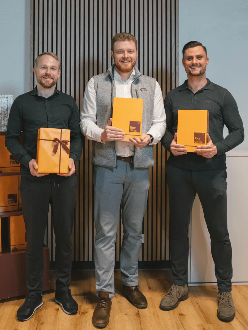 Three men holding orange gift boxes and smiling, standing indoors.