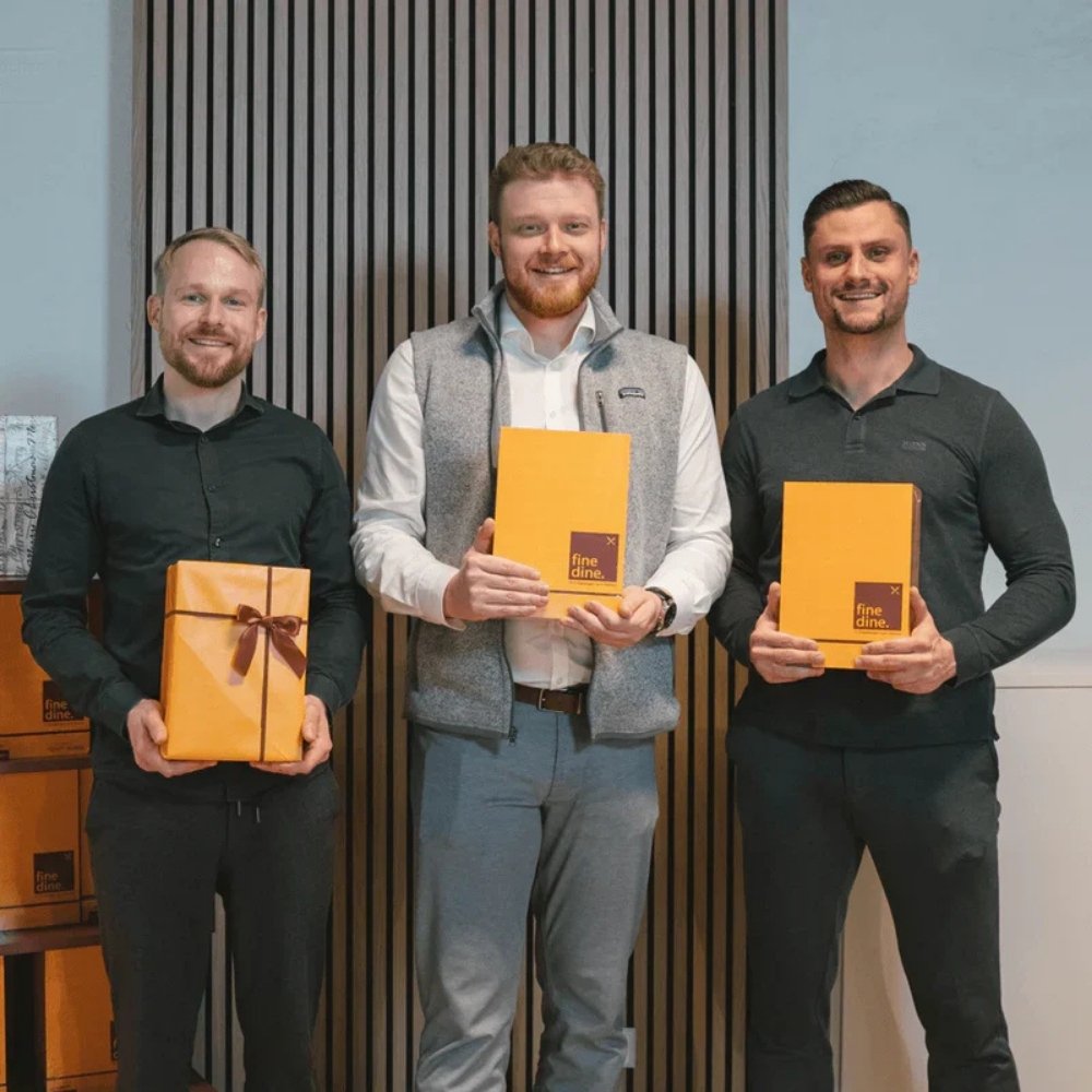 Three men holding orange Fine Dine boxes and gifts, standing in front of a striped wall.