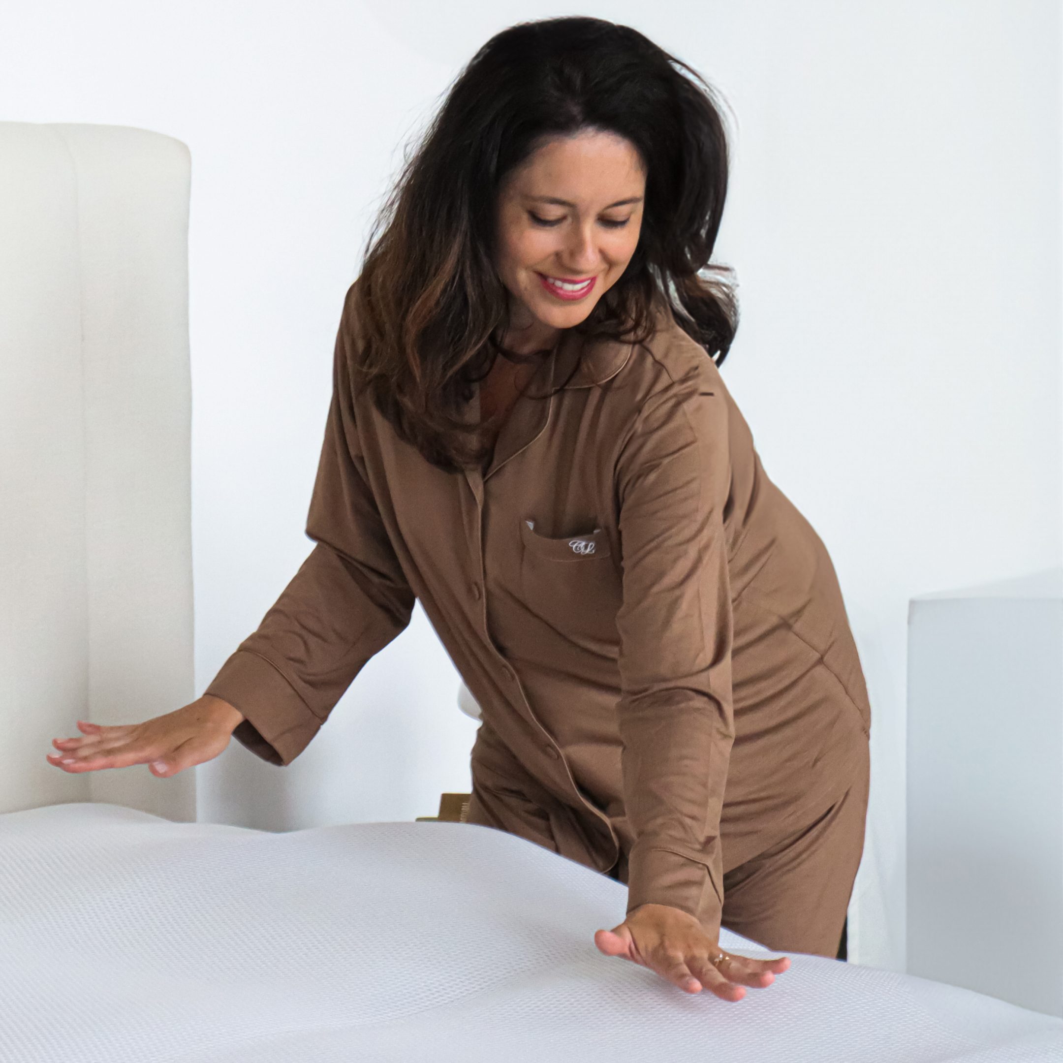 A woman in brown pajamas touching a made bed.