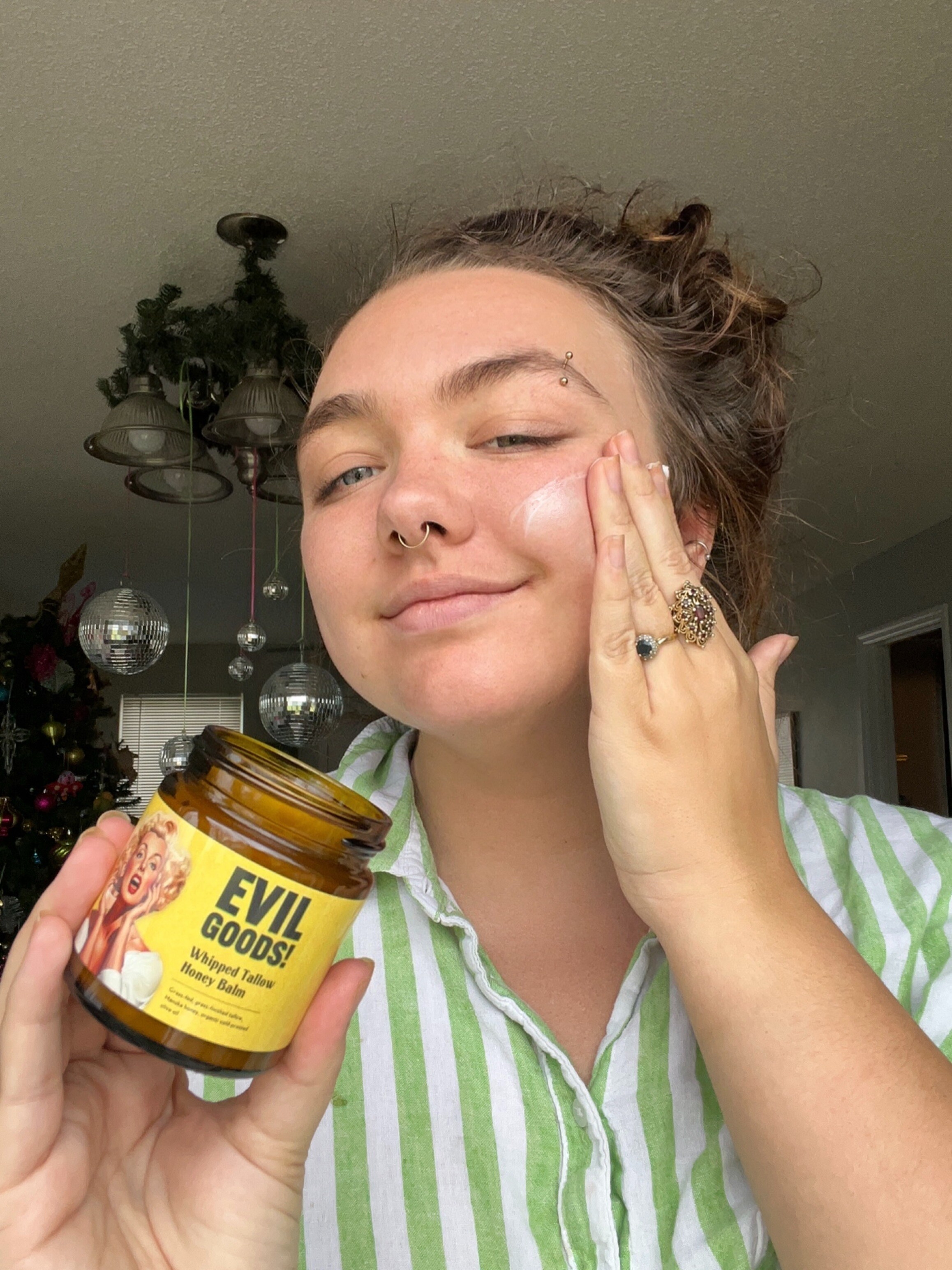 Person applying balm to face, holding a jar labeled 'Evil Goods!' in a home setting.