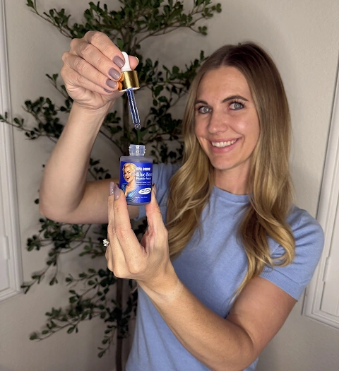 A smiling woman in a blue shirt holds up a bottle of blue serum and a dropper.