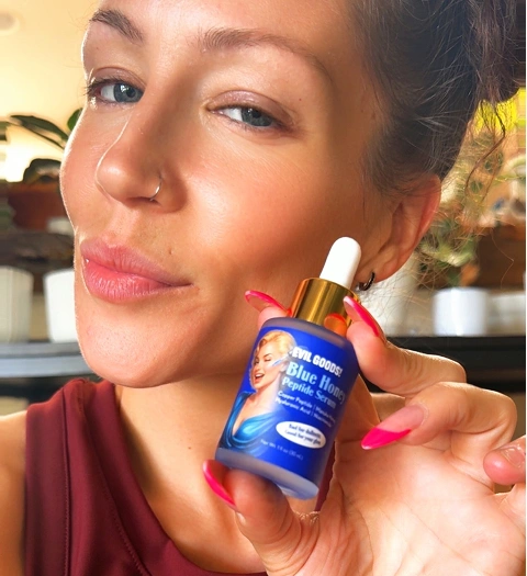 A woman with pink nails holds a small blue bottle of Evil Goods Blue Honey Peptide Serum.