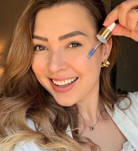 A smiling woman holds a dropper with blue liquid to her cheek, about to apply a skincare serum.